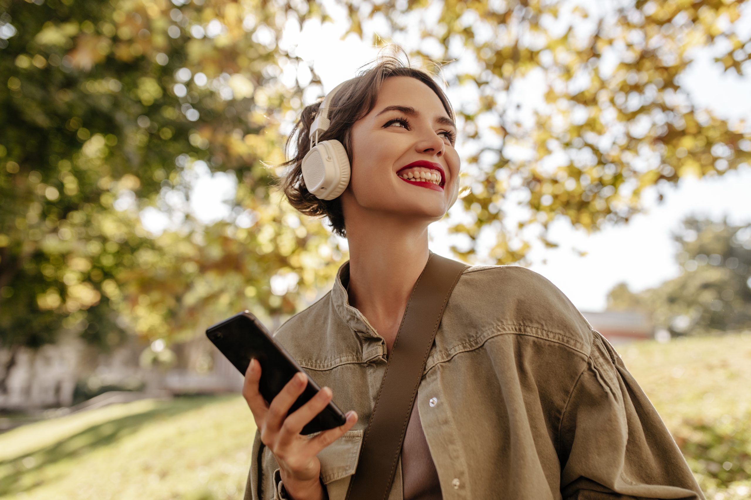 A cheerful woman wearing headphones holds a smartphone while enjoying a sunny day outdoors, symbolizing the positive effects of psychedelic therapy.